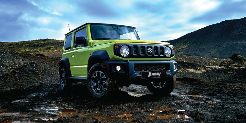Suzuki Jimny verde, un todoterreno compacto, posado en un camino de tierra con montañas de fondo.